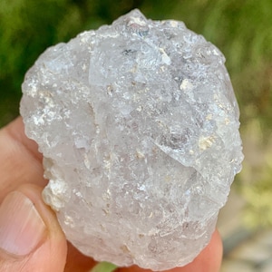 Può includere: Un gruppo di cristalli di quarzo trasparente dalla consistenza ruvida. Il cristallo ha un aspetto traslucido con strutture interne visibili e una lucentezza leggermente iridescente. La formazione naturale è tenuta in mano su uno sfondo verde sfocato.