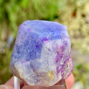 May include: A raw, uncut gemstone with a blend of purple, lavender, and white hues. The crystal has a rough, textured surface and is displayed on a clear stand. The natural stone showcases a unique color gradient.