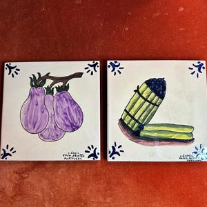 May include: Two square ceramic tiles with hand-painted designs. One tile features three purple eggplants on a branch. The other tile depicts a bundle of asparagus. Both tiles have blue corner accents and text that reads "Hand-Printed Portugal."