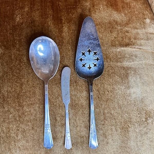 May include: A set of three vintage silver serving utensils: a large spoon, a butter knife, and a pie server. The spoon has a rounded bowl. The pie server has a decorative cut-out design. All pieces have detailed handle designs.