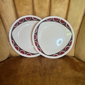 May include: Two white ceramic plates with a maroon and brown geometric design around the rim. The plates are round with a curved design on the side. The plates are on a gold-colored velvet chair.