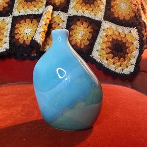 May include: A small, teardrop-shaped ceramic vase with a glossy, light blue finish. The vase has a smooth, curved body with a narrow neck. The background features a colorful, crocheted blanket.