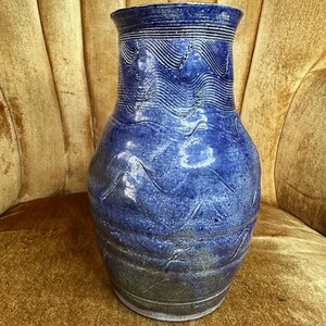 May include: A handcrafted blue ceramic vase with a textured, wave-like pattern. The vase has a wide body and a narrower neck, with horizontal lines near the rim. The vase is approximately 12 inches tall.