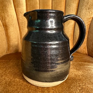May include: A black ceramic pitcher with a brown glaze and a white rim. The pitcher has a rounded body and a handle.