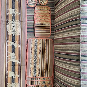 May include: A collection of woven textile items with a Southwestern design. The set includes a runner, placemats, and coasters. The runner and placemats feature a repeating pattern of llamas and geometric shapes in shades of brown, orange, and black, with a pink border.