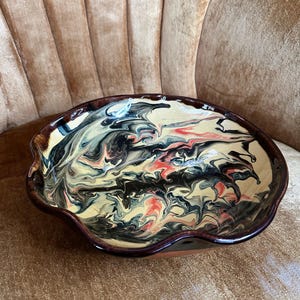 May include: A ceramic bowl with a unique, swirled design. The interior features a mix of black, cream, and red colors, creating an abstract pattern. The bowl has a dark brown rim and a wavy edge, adding to its artistic appeal.