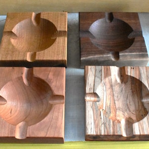 May include: Four wooden square ashtrays with a round, slightly indented center and four small grooves around the center. The ashtrays are made of different types of wood, including cherry, walnut, and maple.