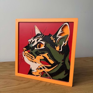 Puede incluir: Un recorte de madera enmarcado de la cabeza de un gato con un esquema de color negro, verde y naranja. El gato mira hacia la derecha y tiene una mancha blanca en el pecho. El marco es naranja.