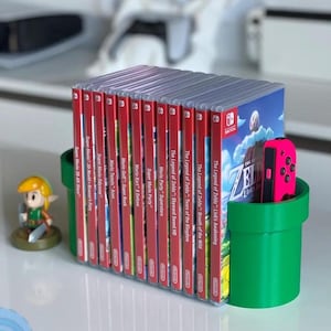 Puede incluir: Un soporte de plástico verde y negro para guardar cartuchos de juegos de Nintendo Switch. El soporte tiene forma de tubo verde y tiene una ranura para un mando Joy-Con de Nintendo Switch. El soporte contiene una variedad de cartuchos de juegos de Nintendo Switch, incluyendo "The Legend of Zelda: Breath of the Wild", "The Legend of Zelda: Tears of the Kingdom" y "Super Mario Odyssey".