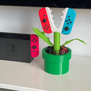以下が含まれることがあります： スーパーマリオブラザーズビデオゲームシリーズのピラニアプラントの形をした、3Dプリントされたニンテンドースイッチゲームカートリッジホルダー。ホルダーは緑色のベース、緑色の茎、そしてニンテンドースイッチ用の赤と青のジョイコンコントローラーを備えています。