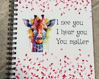 Puede incluir: Un cuaderno de espiral con fondo blanco y una ilustración colorida de una jirafa. El cuaderno tiene confeti rosa y rojo esparcido por la portada. El texto "I see you I hear you You matter" está impreso en la portada.