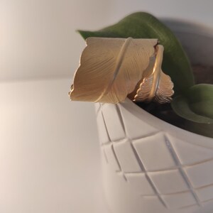 Può includere: Set di anelli a forma di piuma dorati in un vaso di ceramica bianca con motivo a griglia.