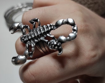 May include: A silver-colored scorpion ring. The ring features a detailed scorpion design with articulated legs, a segmented body, and a curved tail. The scorpion's claws are open, and the ring is worn on a finger.