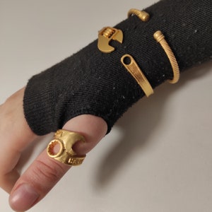 Puede incluir: Un anillo de color dorado en forma de llave inglesa en un dedo, con tres pulseras doradas en la muñeca. Las pulseras son una banda simple, una banda más gruesa con una superficie texturizada y un diseño en forma de llave inglesa.