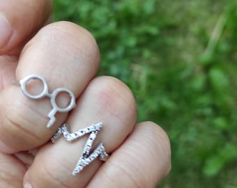 May include: Two silver rings are displayed on a hand. One ring is shaped like a pair of glasses with a lightning bolt, and the other is a zig-zag design with small, clear stones. The background is blurred green.