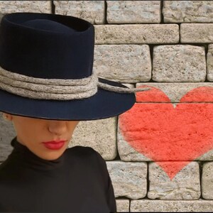 May include: A woman wearing a wide-brimmed navy blue hat with a thick gray rope band and a black turtleneck shirt. A red heart is painted on a brick wall behind her.
