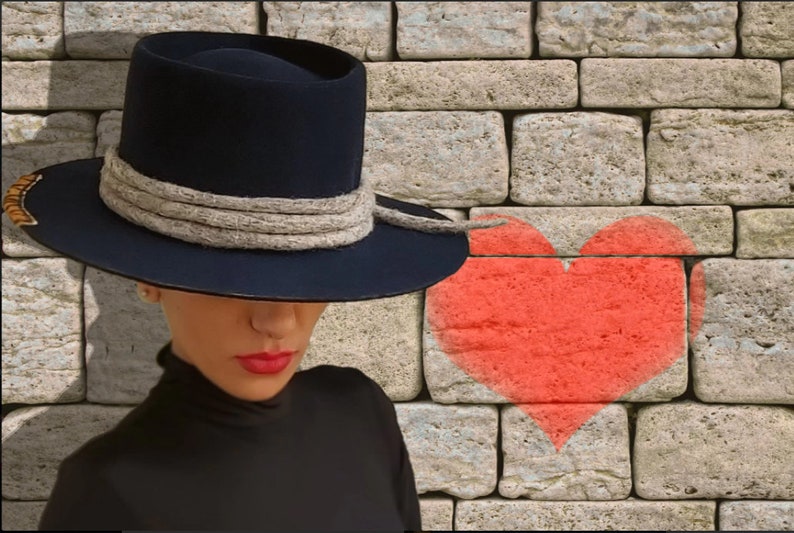 May include: A woman wearing a wide-brimmed navy blue hat with a thick gray rope band and a black turtleneck shirt. A red heart is painted on a brick wall behind her.