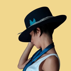 May include: A person wearing a black wide-brimmed hat with a blue feather accent and a blue scarf. The hat has a grey band around the crown.