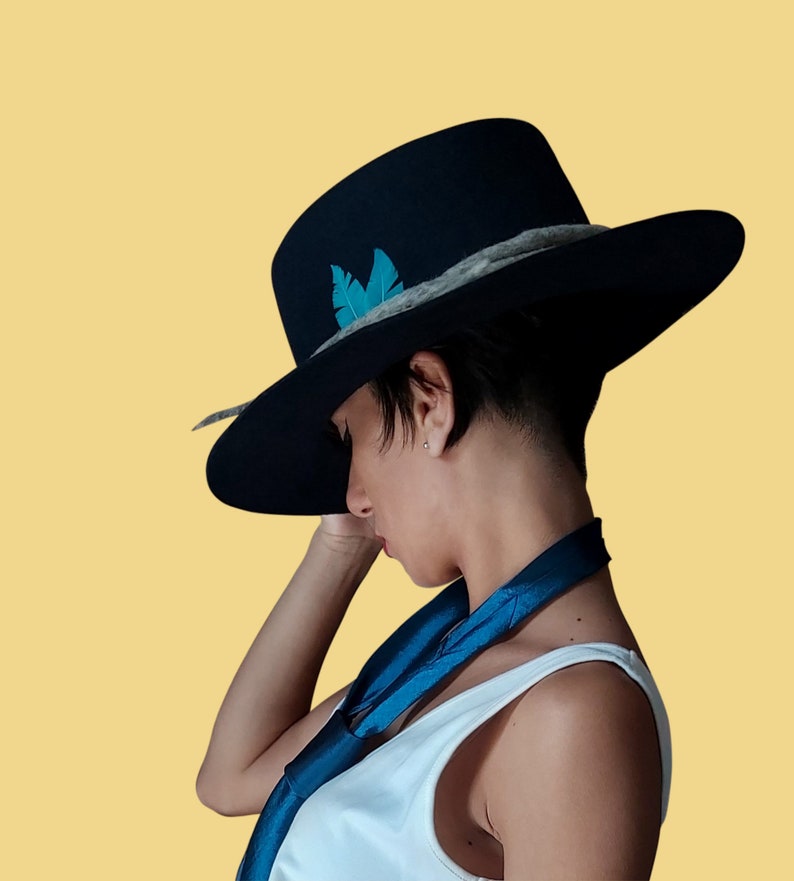 May include: A person wearing a black wide-brimmed hat with a blue feather accent and a blue scarf. The hat has a grey band around the crown.