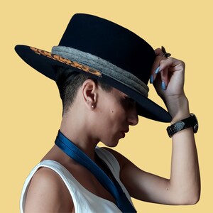 May include: A person wearing a black fedora hat with a leopard print band and a grey ribbon. The hat has a wide brim and is tilted slightly to the side. The person is wearing a white tank top and has their arm raised.
