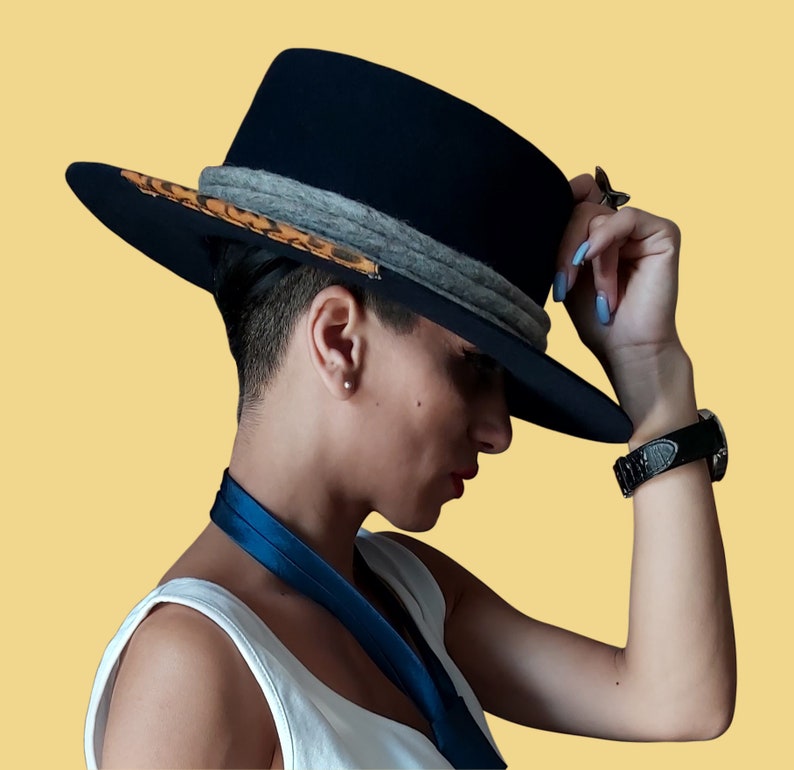 May include: A person wearing a black fedora hat with a leopard print band and a grey ribbon. The hat has a wide brim and is tilted slightly to the side. The person is wearing a white tank top and has their arm raised.