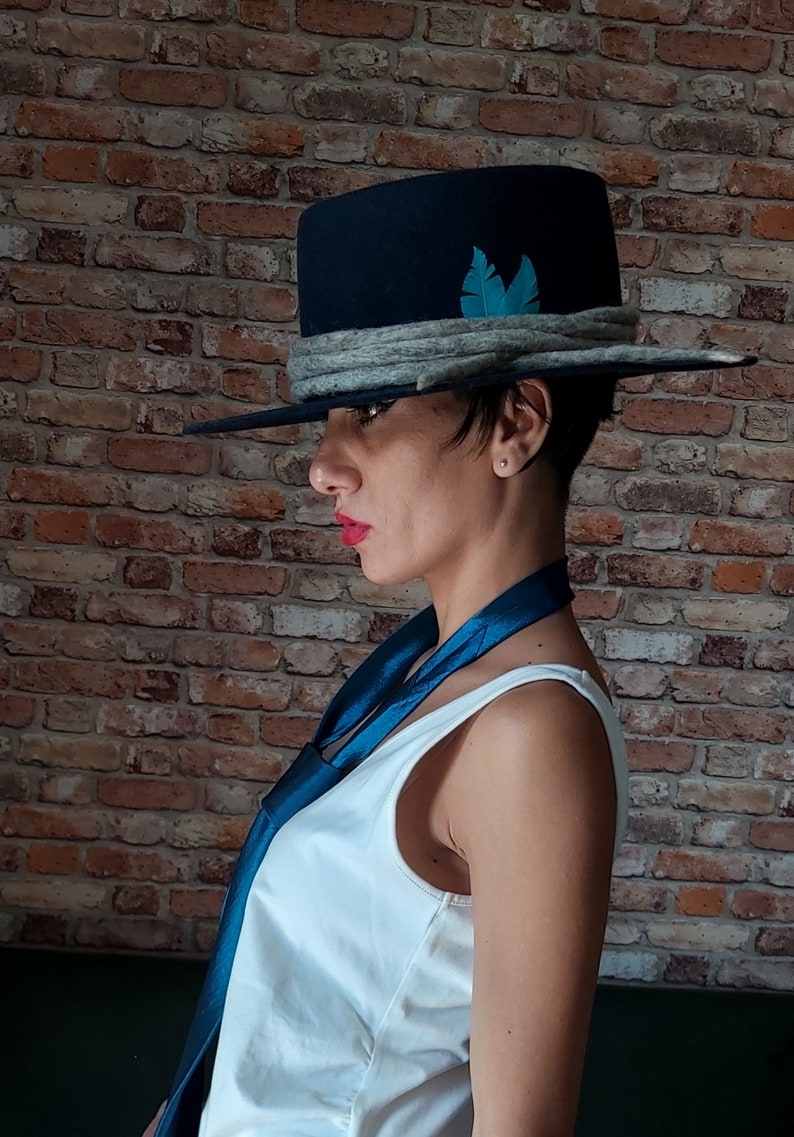 May include: A woman wearing a black fedora with a gray band and a teal feather accent. She is also wearing a white tank top and a teal blue scarf.