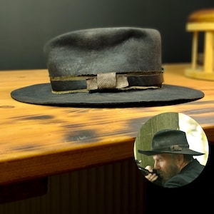 Pode incluir: Um chapéu fedora de feltro cinza escuro com aba larga e uma faixa de couro com uma fivela decorativa. O chapéu tem um aspecto envelhecido e está sobre uma superfície de madeira.