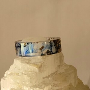 May include: A resin ring with a marbled blue and white design, accented with gold leaf flakes. The ring is displayed on a white, crystalline surface, showcasing its unique, artistic style. This handcrafted ring is a statement piece.