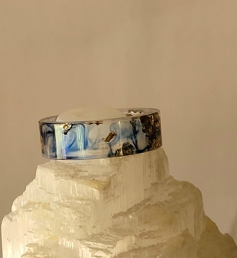 May include: A resin ring with a marbled blue and white design, accented with gold leaf flakes. The ring is displayed on a white, crystalline surface, showcasing its unique, artistic style. This handcrafted ring is a statement piece.