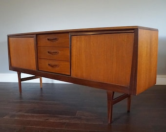May include: A mid-century modern teak wood sideboard with three drawers and two doors. The sideboard has a light wood finish and is supported by four legs.