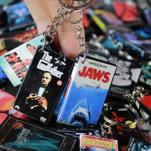 Può includere: Due portachiavi rettangolari con miniature delle copertine dei film The Godfather e Jaws. Ogni portachiavi è fissato a una catena e un anello in metallo argentato. La copertina di The Godfather è nera con testo bianco, Jaws è bianca e blu.