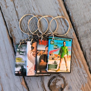 May include: Keyrings featuring miniature DVD covers from the TV show "Breaking Bad." Each keyring displays a different season's cover art, with vibrant colours and recognisable imagery. The keyrings are attached to silver metal rings.