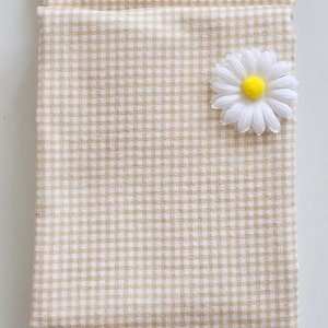 May include: A folded beige and white gingham fabric with a white daisy flower with a yellow center. The fabric is a light tan color and has a classic checkered pattern. The daisy is placed on the upper right corner.