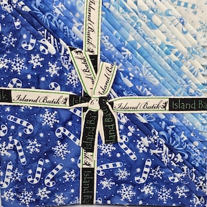 May include: A bundle of 12 fat quarter fabric squares featuring a blue and white winter-themed print with snowflakes and candy canes. The fabric is tied with a ribbon that reads "Island Batik".