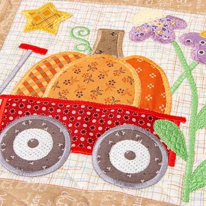 May include: A quilted patch featuring a red wagon carrying a pumpkin with orange and yellow patterns. The wagon has brown wheels and is decorated with a yellow star and purple flowers. The background is a beige fabric with printed text.