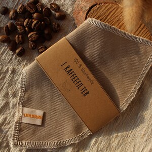 Könnte beinhalten: Ein beigefarbener wiederverwendbarer Kaffeefilter aus 100% Baumwolle mit einem braunen Papierlabel mit der Aufschrift "1 KAFFEEFILTER 100% Baumwolle".