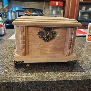 Pode incluir: Uma caixa de joias de madeira castanha clara com tampa articulada e fecho de metal ornamentado. A caixa tem entalhes decorativos nas laterais e assenta em pequenos pés decorativos. O fundo mostra um ambiente de cozinha.