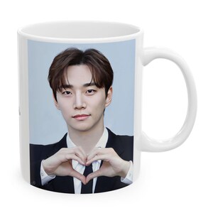 May include: A white ceramic mug featuring a photographic print of a person with dark brown hair, wearing a black suit jacket, white dress shirt, and black tie, using both hands to form a heart shape in front of their chest against a light background.