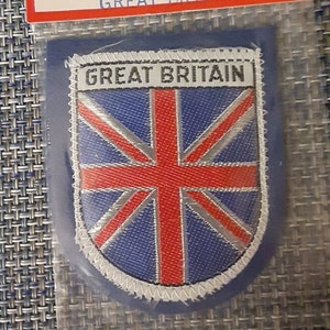 May include: A patch featuring the flag of Great Britain, with a red cross and white and blue background. The patch is in a shield shape and is labeled "GREAT BRITAIN". The packaging reads "AMERICAN HOUSE OF FLAGS PRODUCT".