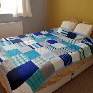 Pu&ograve; includere: Un trapunta patchwork blu, grigia e bianca realizzata con quadrati di tessuto a maglia. La trapunta &egrave; su un letto con una testata bianca e un copriletto con motivo dorato.