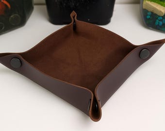 May include: A dark brown leather tray, designed to catch small items. The tray has a square shape with snap closures at each corner. The interior is a lighter brown, suede-like material. The corners are folded up to form the tray.