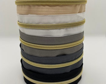 May include: A stack of five zipper tapes with gold metal teeth. The tapes are in various colors, including beige, white, gray, and black.