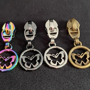 Könnte beinhalten: Vier Reißverschluss-Zipper mit Schmetterlingsanhängern. Die Anhänger sind kreisförmig mit einem Schmetterlingsausschnitt. Die Reißverschluss-Zipper sind in Regenbogen, Silber, Gold und Bronze.