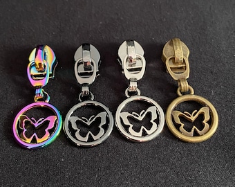 Può includere: Quattro tiretti per cerniera con ciondoli a forma di farfalla. I ciondoli sono di forma circolare con un ritaglio a forma di farfalla. I tiretti per cerniera sono in arcobaleno, argento, oro e bronzo.