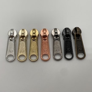 Könnte beinhalten: Sieben Metall-Reißverschluss-Schieber in verschiedenen Farben: Silber, Gold, Bronze, Kupfer, Gunmetal, Schwarz und Antik-Messing. Jeder Schieber hat eine abgerundete Oberseite und eine kreisförmige Öffnung unten. Die Schieber sind in einer Reihe angeordnet.