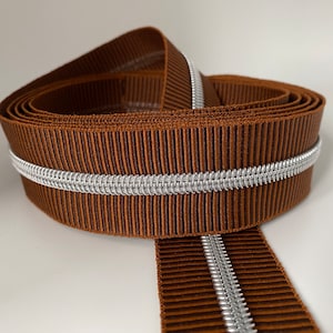 Könnte beinhalten: Braunes Band mit silbernem Metallic-Besatz. Das Band ist gefaltet und zeigt den Besatz auf beiden Seiten.