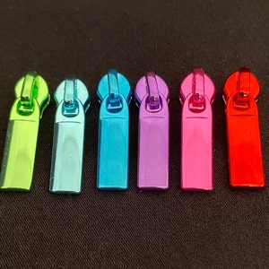 Könnte beinhalten: Sechs Reißverschluss-Zipper in verschiedenen Farben: Grün, Türkis, Blau, Lila, Pink und Rot. Die Reißverschluss-Zipper sind alle gleich groß und haben die gleiche Form, und sie sind alle aus Metall.
