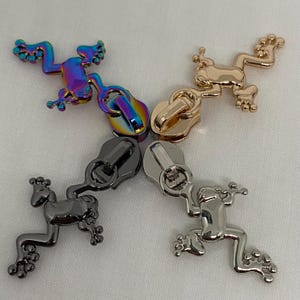 Könnte beinhalten: Vier Metall-Reißverschlussanhänger in Form von Fröschen. Einer ist schillernd, einer goldfarben, einer anthrazitfarben und einer silberfarben. Die Frosch-Anhänger befinden sich auf weißem Hintergrund.