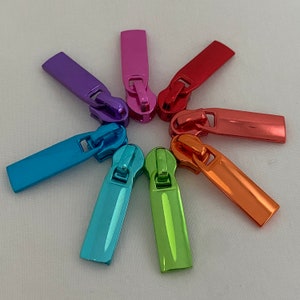 Könnte beinhalten: Ein Satz von acht Reißverschluss-Zierbändern in verschiedenen Metallic-Farben, darunter Blau, Grün, Orange, Rot, Pink und Lila. Die Bänder sind sternförmig auf einem weißen Hintergrund angeordnet.
