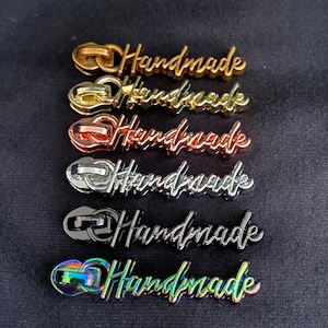 Könnte beinhalten: Sechs Reißverschluss-Zipper mit dem Wort "Handmade" in einer kursiven Schrift. Die Zipper sind in verschiedenen Farben: Gold, Silber, Roségold, Silber, Schwarz und schillernd.
